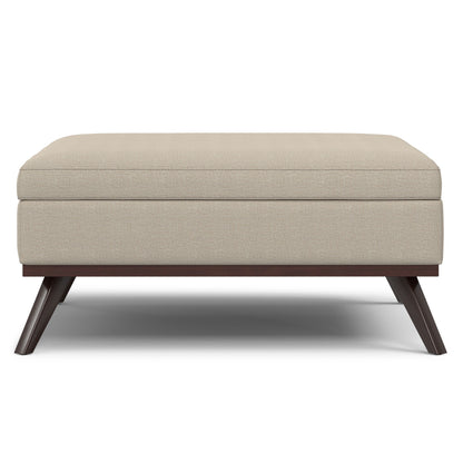 The Owen Multifunctional Square Coffee Table Storage Ottoman features beige upholstery, a cushioned top, and angled dark wooden legs, all presented against a white background.