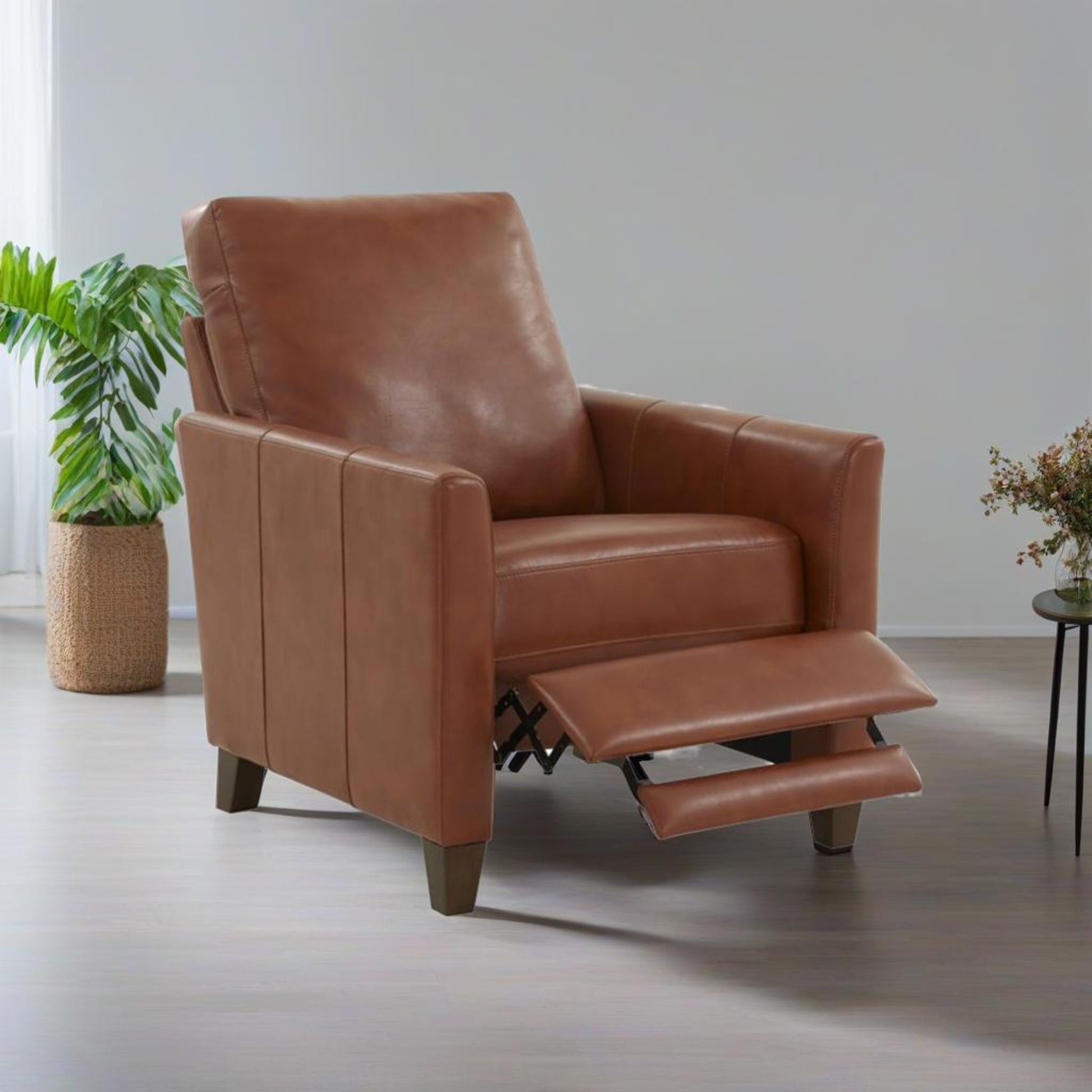 The Pablo - Modern Recliner With Manual Recline, in brown leather with its footrest extended, stands on a light wood floor near a green potted plant and a small table with flowers against a plain light wall.