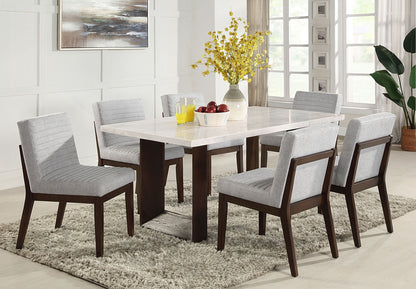 A modern dining room features the Edwyn Marble Top Dining Table - Natural, six gray upholstered chairs, a beige rug, and a yellow flower centerpiece. A bowl of apples and a jug of orange juice sit on the table below large sunlit windows.