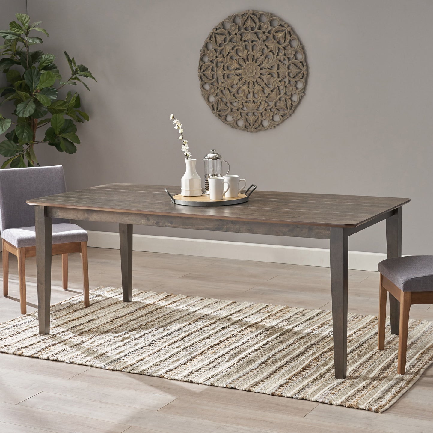 The Farmhouse Charm Dining Table With Square Leg and two chairs are placed on a woven rug, set with a tray, teapot, two cups, and a vase of white flowers. A decorative wall piece hangs above and a potted plant is in the corner.