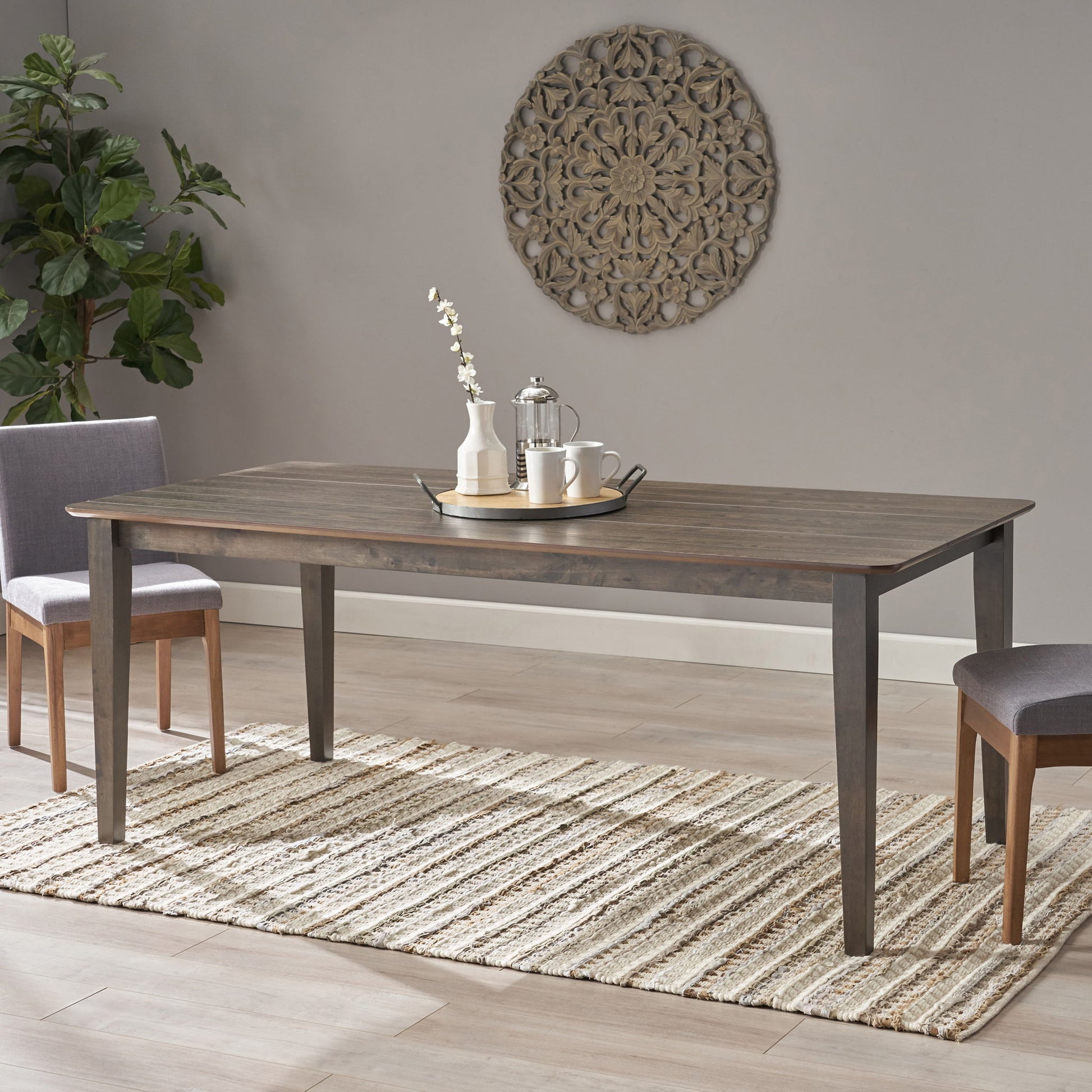 The Farmhouse Charm Dining Table With Square Leg and two chairs are placed on a woven rug, set with a tray, teapot, two cups, and a vase of white flowers. A decorative wall piece hangs above and a potted plant is in the corner.
