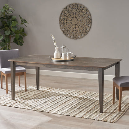 The Farmhouse Charm Dining Table With Square Leg and two chairs are placed on a woven rug, set with a tray, teapot, two cups, and a vase of white flowers. A decorative wall piece hangs above and a potted plant is in the corner.