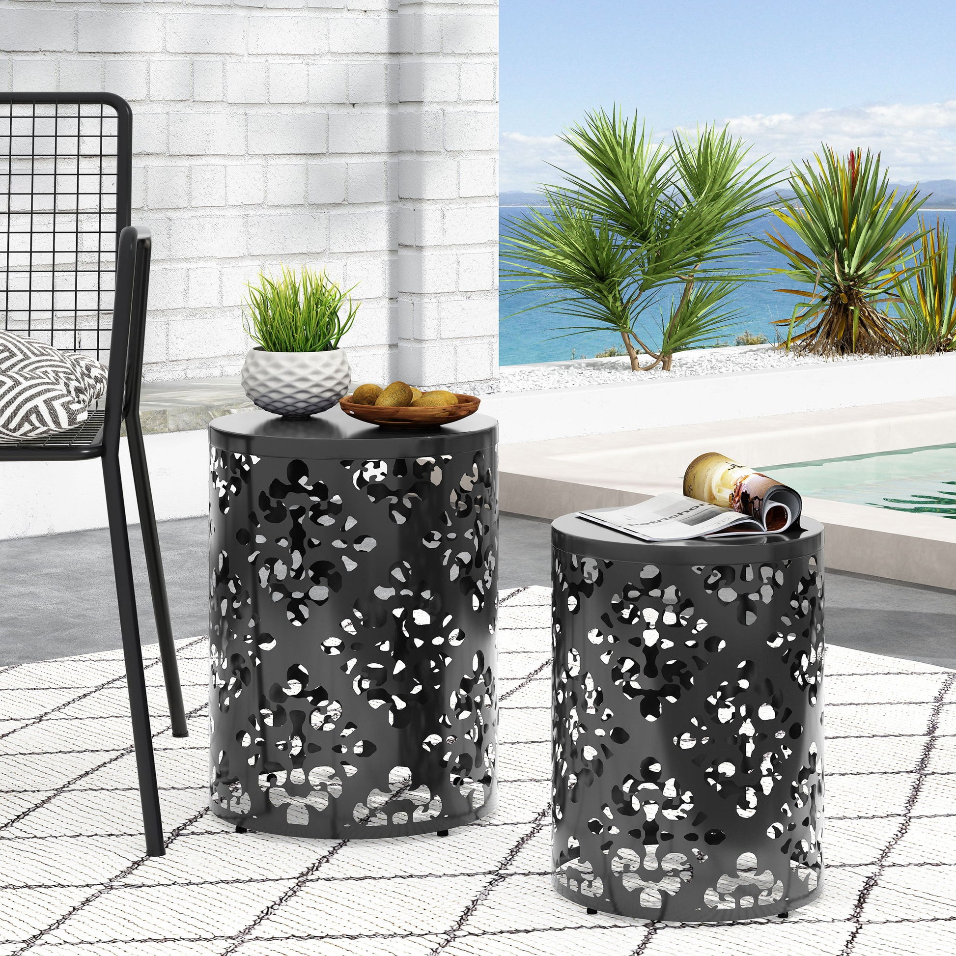 Two Outdoor Metal Modern Design End Tables with cut-out patterns rest on a white patterned rug by the pool—one holds a plant and snacks, the other magazines. Palm trees and ocean provide a scenic backdrop.