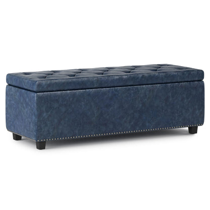 The Hamilton Upholstered Storage Ottoman features a rectangular blue design with a tufted lid, decorative nailhead trim at the base, and short black legs.