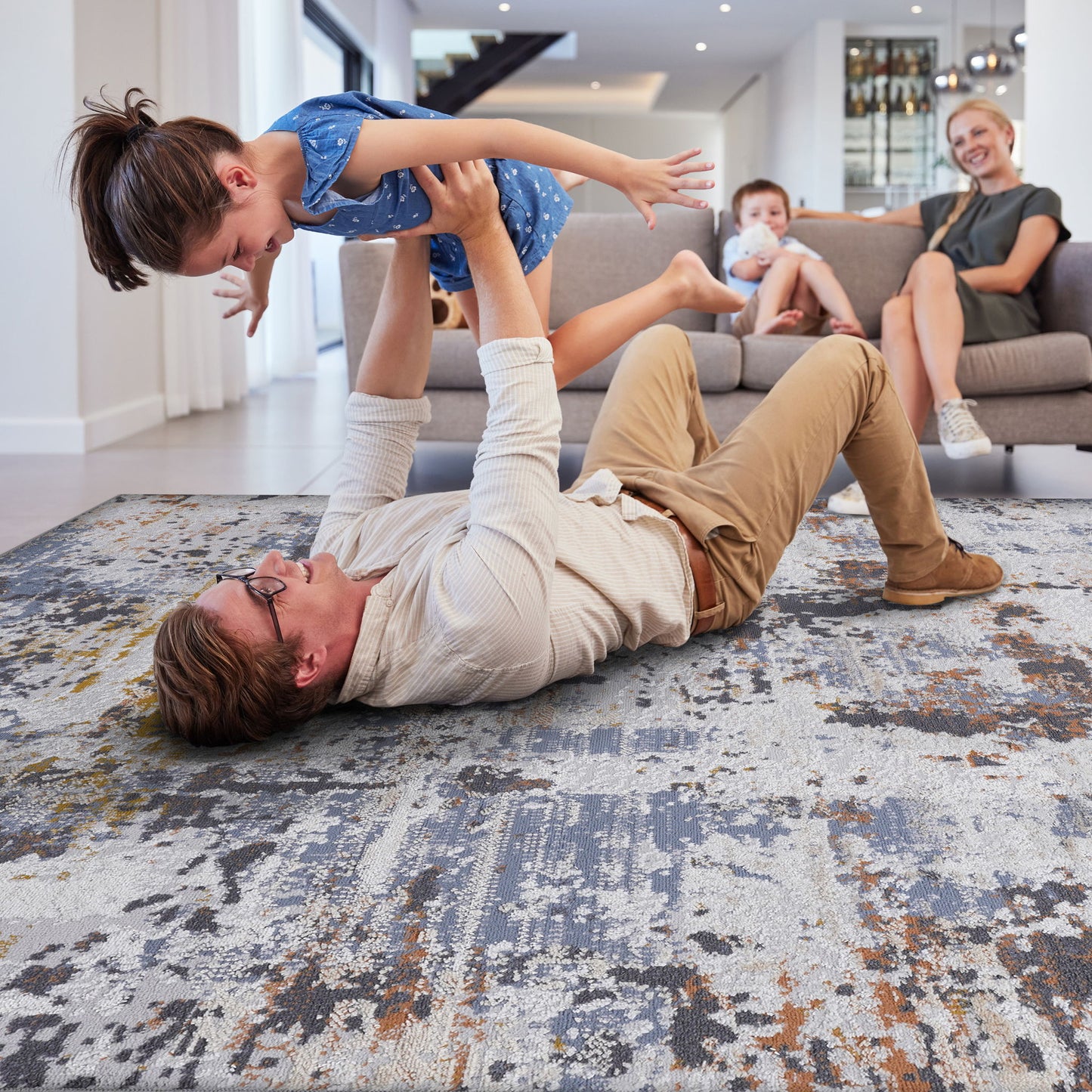 A man lies on the Payas - Abstract Area Rug, lifting a smiling girl in a blue dress above him. In the background, a woman and boy relax on a gray couch, all enjoying time together in a bright living room.