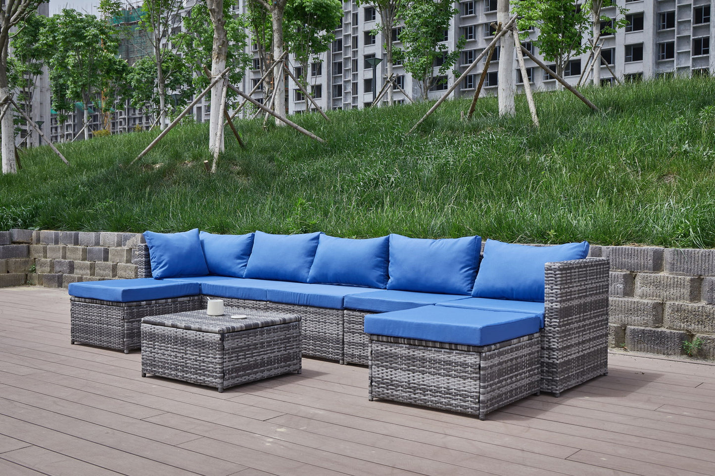 The Patio Sofa With Cushions, featuring grey wicker, blue cushions, a sectional sofa, two ottomans, and a low table, is set on a wooden deck with grass and trees in the background.