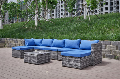 The Patio Sofa With Cushions, featuring grey wicker, blue cushions, a sectional sofa, two ottomans, and a low table, is set on a wooden deck with grass and trees in the background.