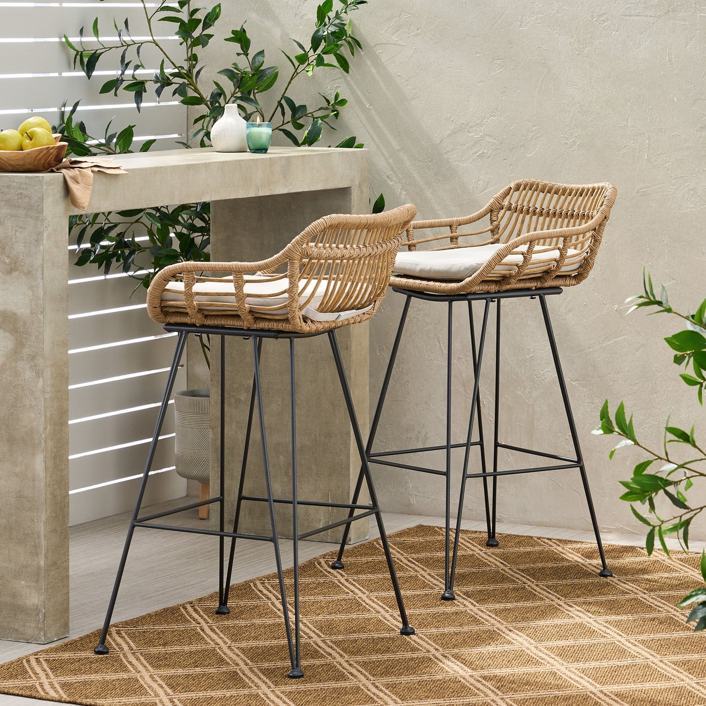Two Outdoor Rattan Bar Chairs With Metal Legs and cushions are placed by a concrete counter in a bright, modern patio with green plants and a woven rug on the floor.