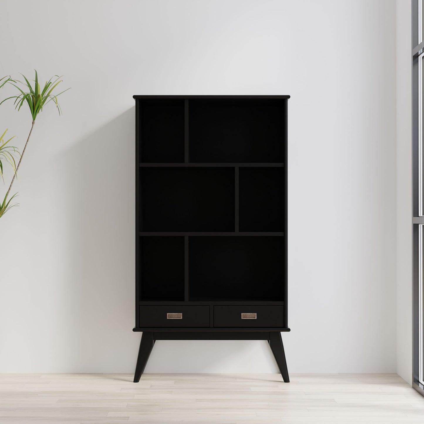 The Draper - Mid Century Wide Bookcase And Storage Unit, featuring open shelves and two bottom drawers in black, stands against a white wall near a window. A green potted plant is to the left on the light wooden floor.