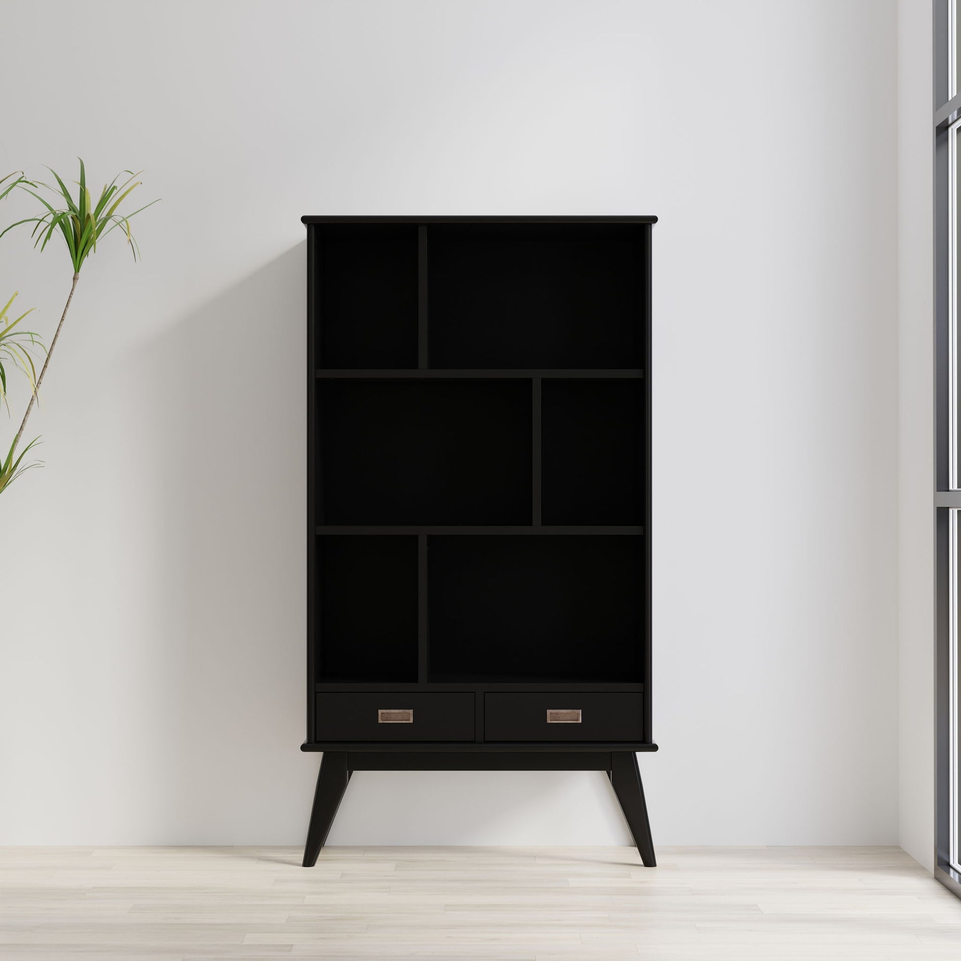 The Draper - Mid Century Wide Bookcase And Storage Unit, featuring open shelves and two bottom drawers in black, stands against a white wall near a window. A green potted plant is to the left on the light wooden floor.