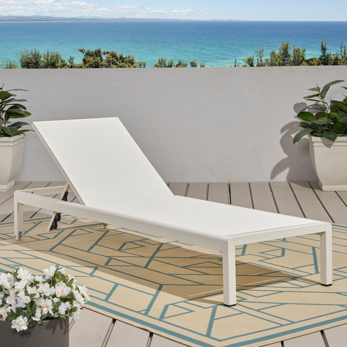 An Outdoor Patio Chaise Lounge With 5 Reclining Positions sits on a patterned rug on a wooden deck with potted plants, set against a white wall and offering a scenic view of the blue ocean and sky.