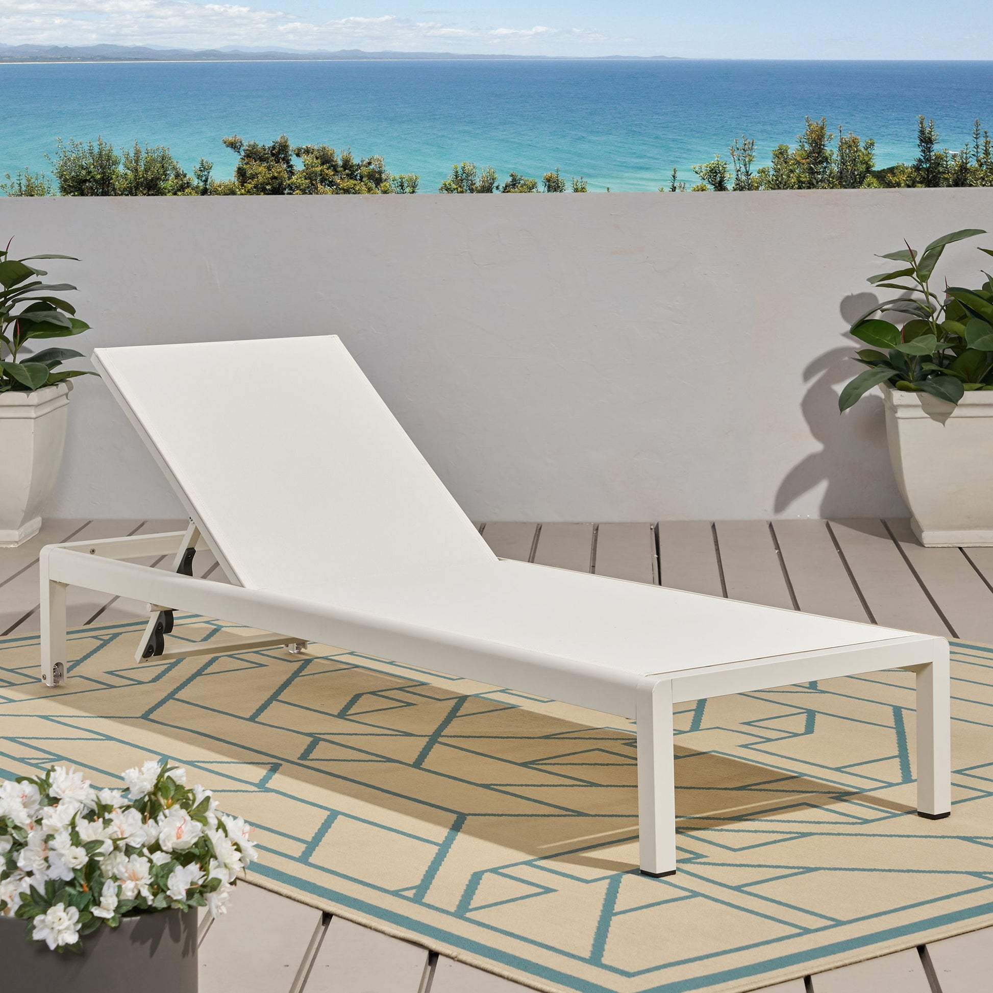 An Outdoor Patio Chaise Lounge With 5 Reclining Positions sits on a patterned rug on a wooden deck with potted plants, set against a white wall and offering a scenic view of the blue ocean and sky.