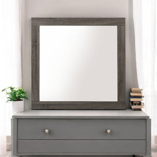 The Avantika Rustic Mirror in gray/oak sits on a gray dresser, with a small potted plant on the left, a stack of books on the right, and sheer white curtains in the background.