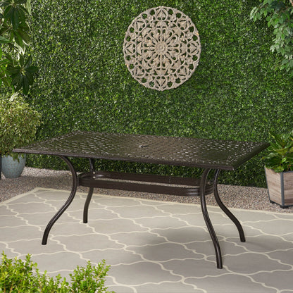 The Celestine Outdoor Cast Aluminum Dining Table with a floral lattice design and umbrella holder sits on a light patterned rug, surrounded by greenery and plants, with a decorative round wall piece on a green ivy wall.