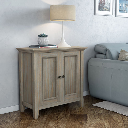 The Amherst - Handcrafted Low Storage Cabinet with two doors stands beside a light gray sofa, topped with a white table lamp, small potted succulent, and closed book. Wall art in the background completes the cozy, modern scene.