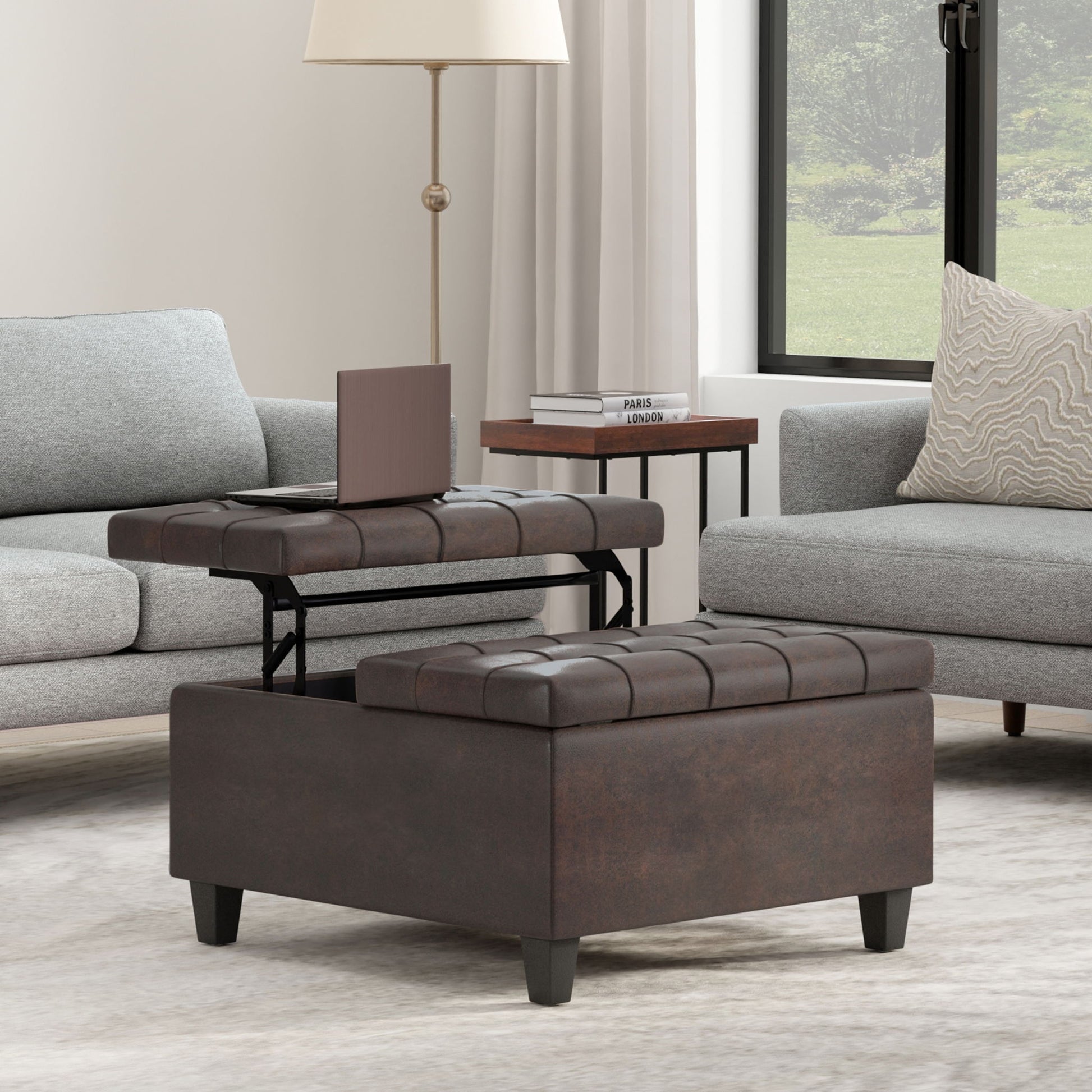 The Harrison Upholstered Coffee Table Storage Ottoman with a lift-top tray sits on a light rug between two gray sofas, with a lamp, books, and a window framed by white curtains in the background.