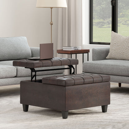 The Harrison Upholstered Coffee Table Storage Ottoman with a lift-top tray sits on a light rug between two gray sofas, with a lamp, books, and a window framed by white curtains in the background.