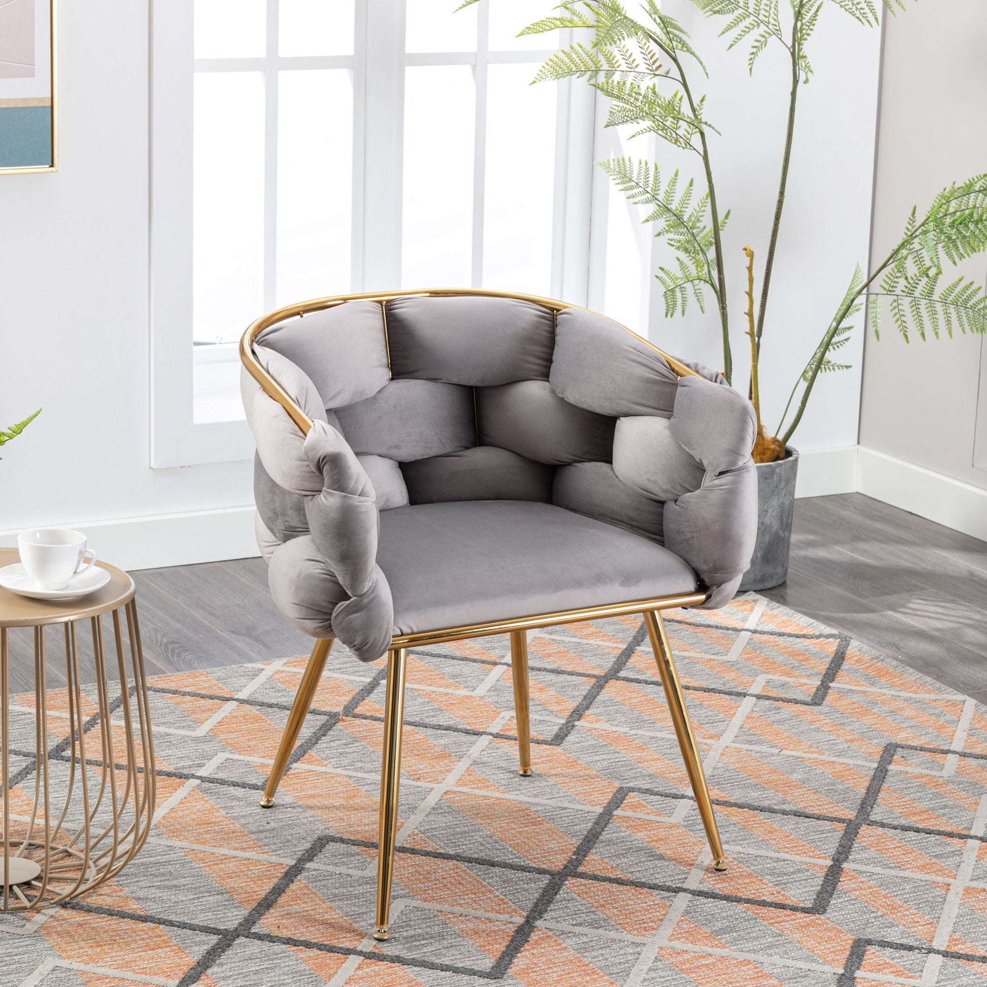 A Luxury Modern Simple Leisure Velvet Single Sofa Chair with gold legs sits on a rug beside a small round table and potted plant in a bright room with large windows.