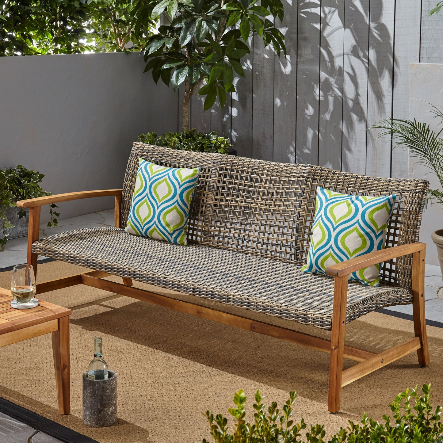 The Outdoor 3 Seater Sofa With Rattan Cover - Gray, styled with two colorful pillows, rests on a tan rug beside a small wooden table holding a glass and bottle, surrounded by potted plants on a sunlit patio.