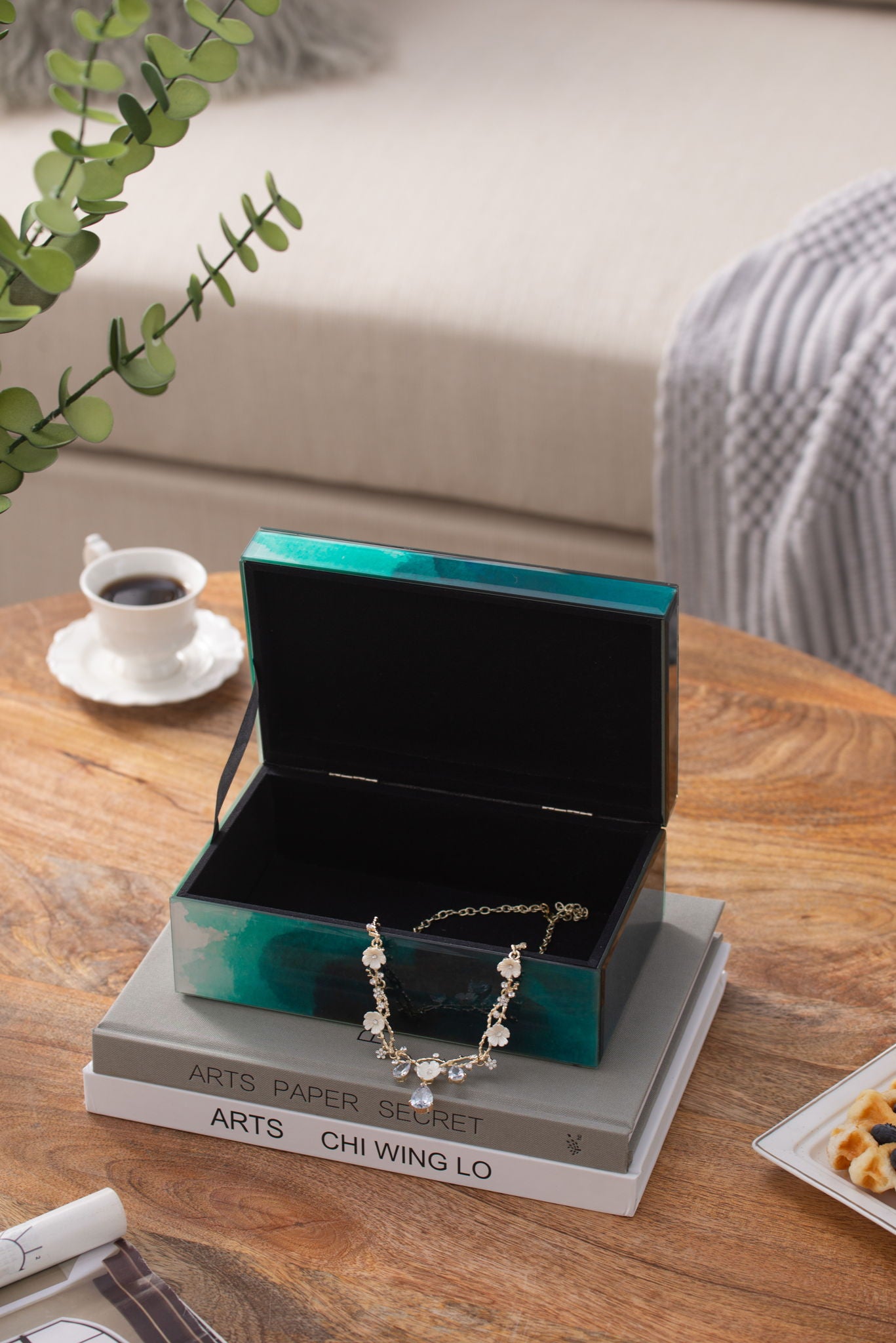 The Bethany - Aqua Jewelry Box with its lid open, displaying a delicate necklace, rests atop art books on a wooden table. Nearby are coffee, waffles, and a plant, with a sofa and blanket in the background.