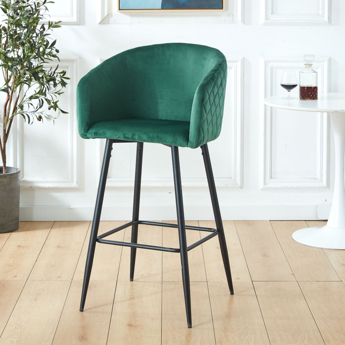 A Counter Height Bar Stool, Metal Legs, Velvet Upholstered Set in Black / Green stands on a light wood floor beside a small white table with a wine glass and decanter, with a leafy potted plant in the background.