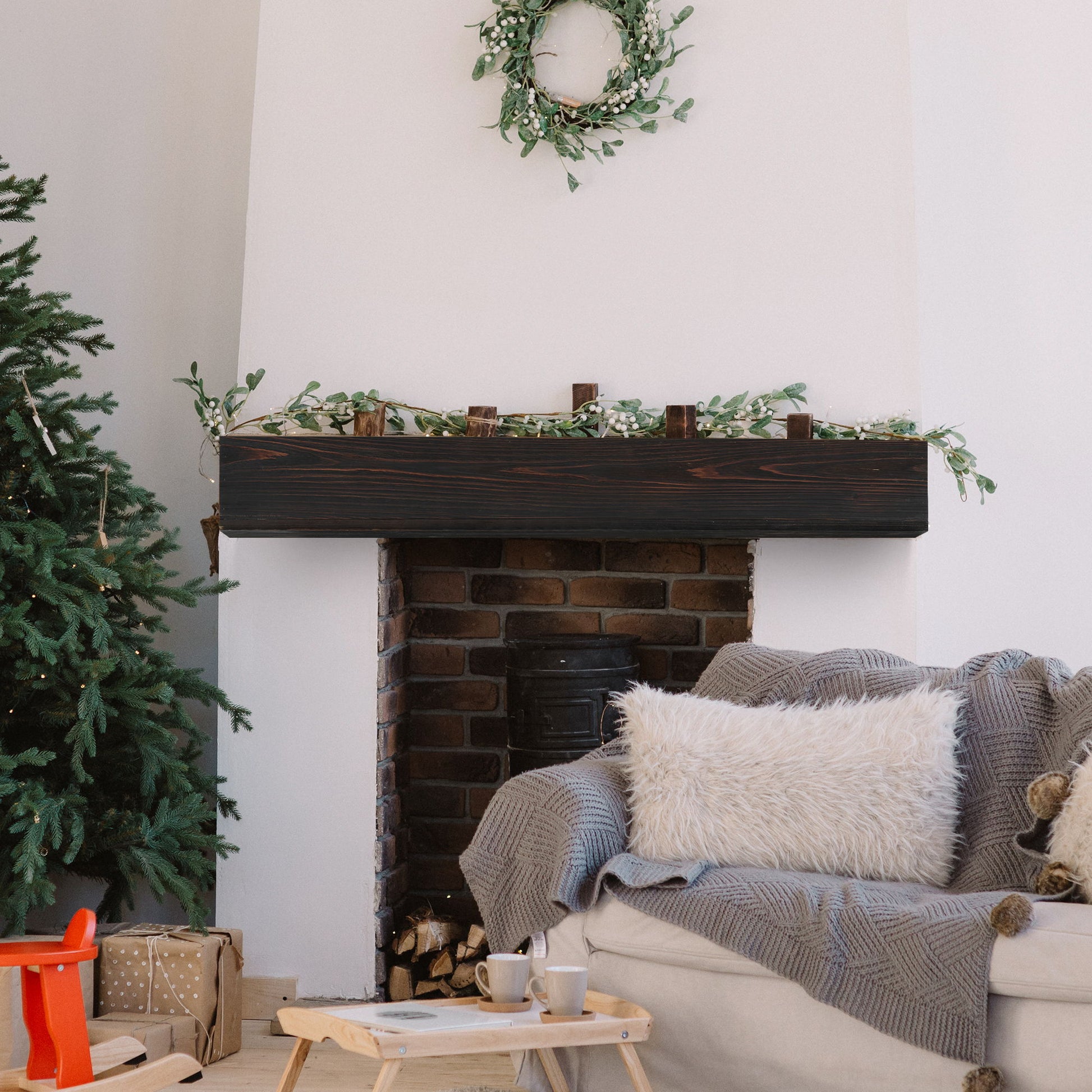 A cozy living room features a decorated Christmas tree, presents, a gray couch with fluffy pillow and blanket, and a fireplace adorned with garland and a wreath above the Rustic Wood Fireplace Mantel floating shelf. A coffee table holds two mugs.