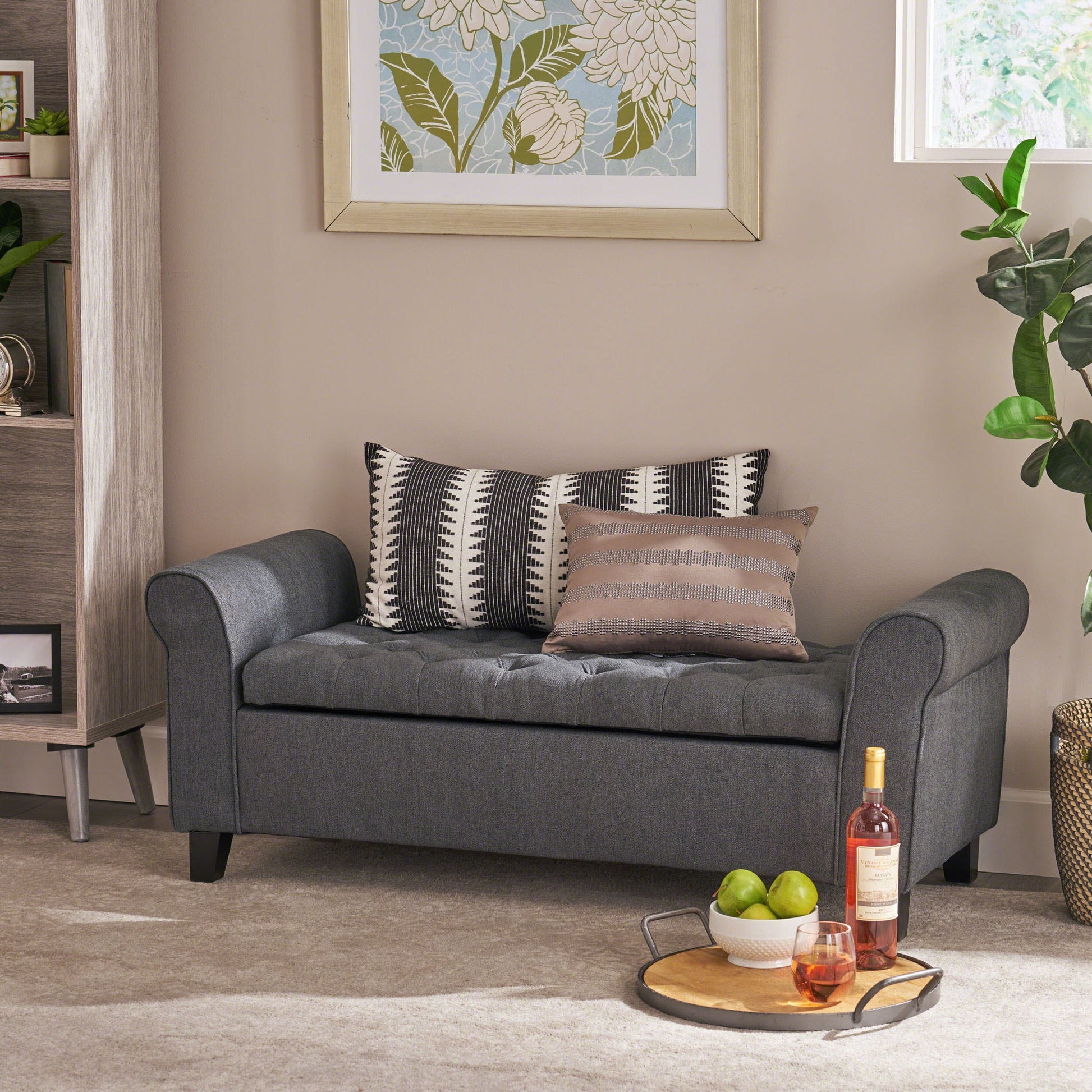 A modern living room showcases the Hayes Contemporary Armed Storage Bench with rolled arms and button tufting, topped with two patterned pillows, a floral artwork above, and a tray holding a wine bottle, glass, and green apples in front.