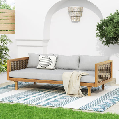 The Claremont 3 Seater Daybed features a teak acacia wood frame, water-resistant light gray cushions, and woven sides. It sits on a blue and white patterned rug with a blanket and pillow, surrounded by green plants against a white wall.