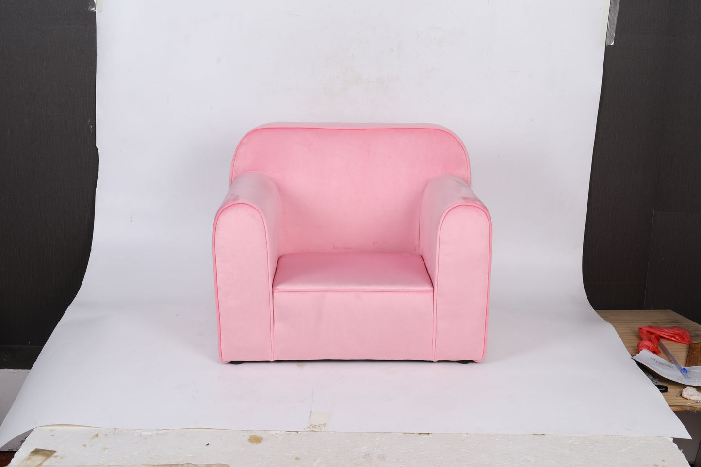 The Durable Design Kids Club Sofa, a small pink armchair with rounded arms and backrest, sits on a white backdrop with some boards and light brown flooring visible at the edges.