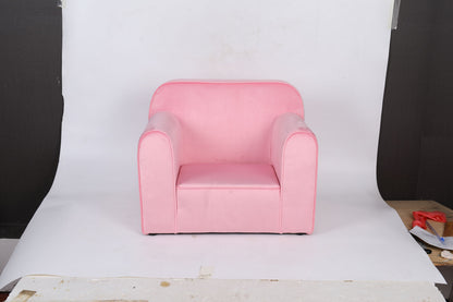 The Durable Design Kids Club Sofa, a small pink armchair with rounded arms and backrest, sits on a white backdrop with some boards and light brown flooring visible at the edges.
