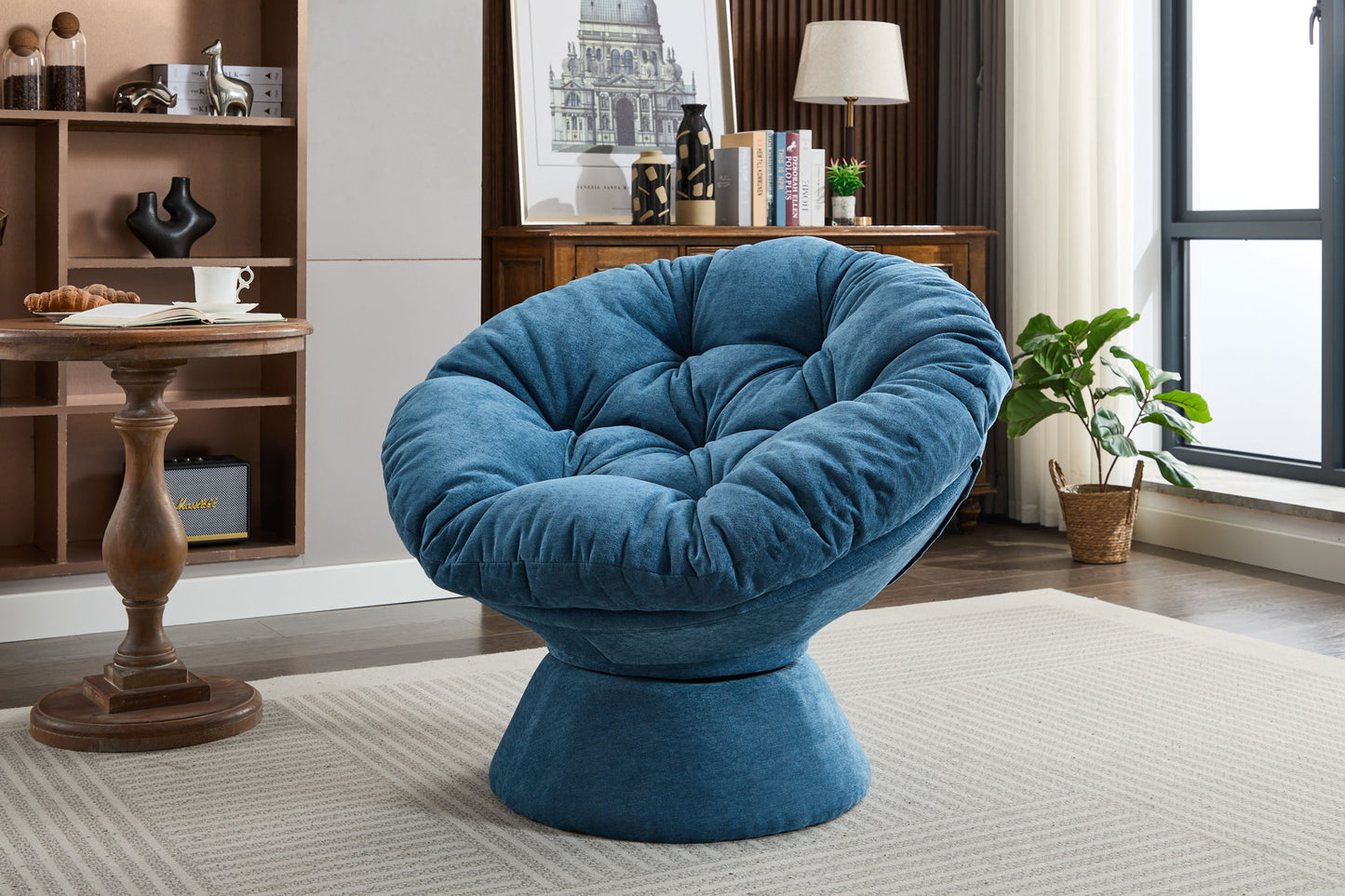 An Oversized Swivel Accent Chair, 360 Swivel Barrel Papasan Chair in blue with tufted cushioning sits on a light patterned rug in a modern living room with bookshelves, a wooden desk, plants, and large windows.