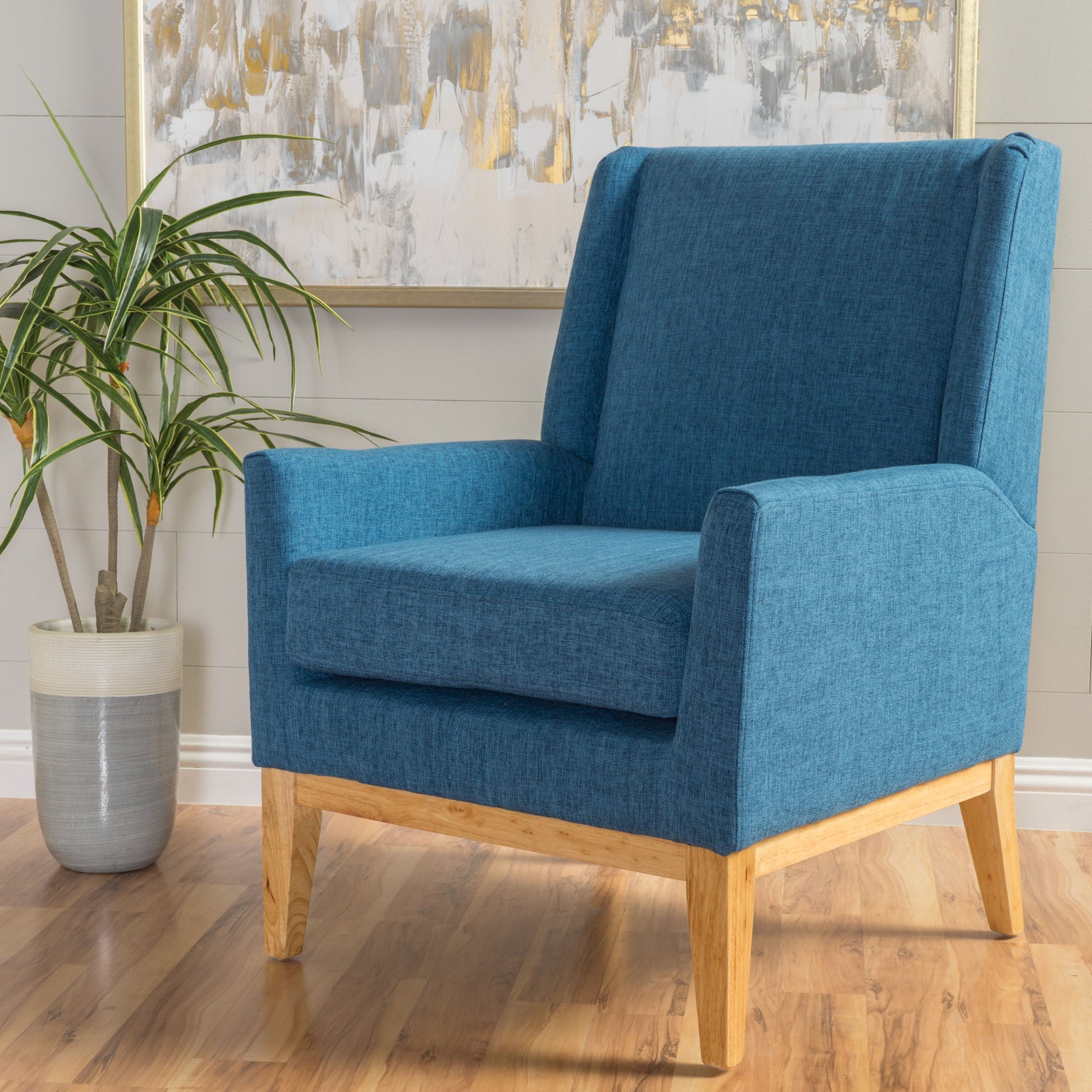 The Modern Accent Chair With Padded Cushion Seat, featuring blue upholstery and wooden legs, is placed on a hardwood floor beside a green potted plant, with an abstract painting on the light gray wall behind.