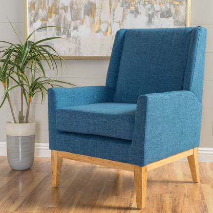 The Modern Accent Chair With Padded Cushion Seat, featuring blue upholstery and wooden legs, is placed on a hardwood floor beside a green potted plant, with an abstract painting on the light gray wall behind.