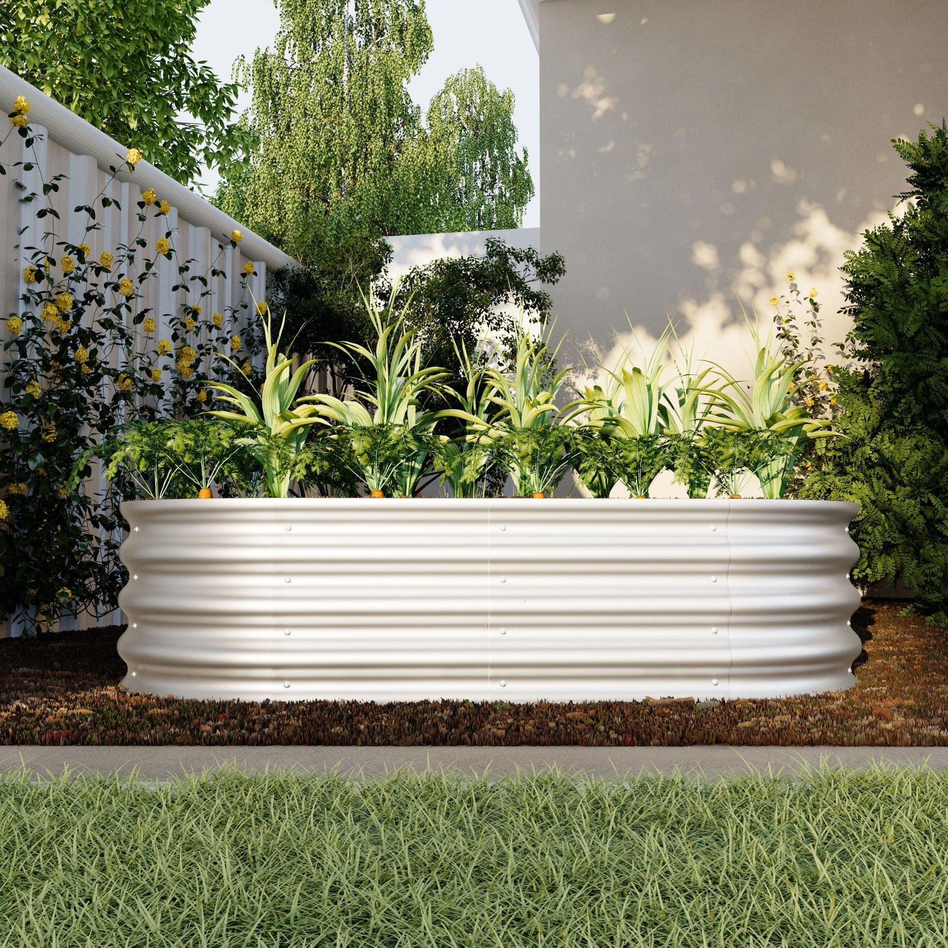 A Heavy Duty Galvanized Raised Garden Bed Kit for vegetables, flowers, and herbs, filled with green plants, sits on brown soil in a backyard surrounded by lawn, trees, bushes, and a white building in the background.