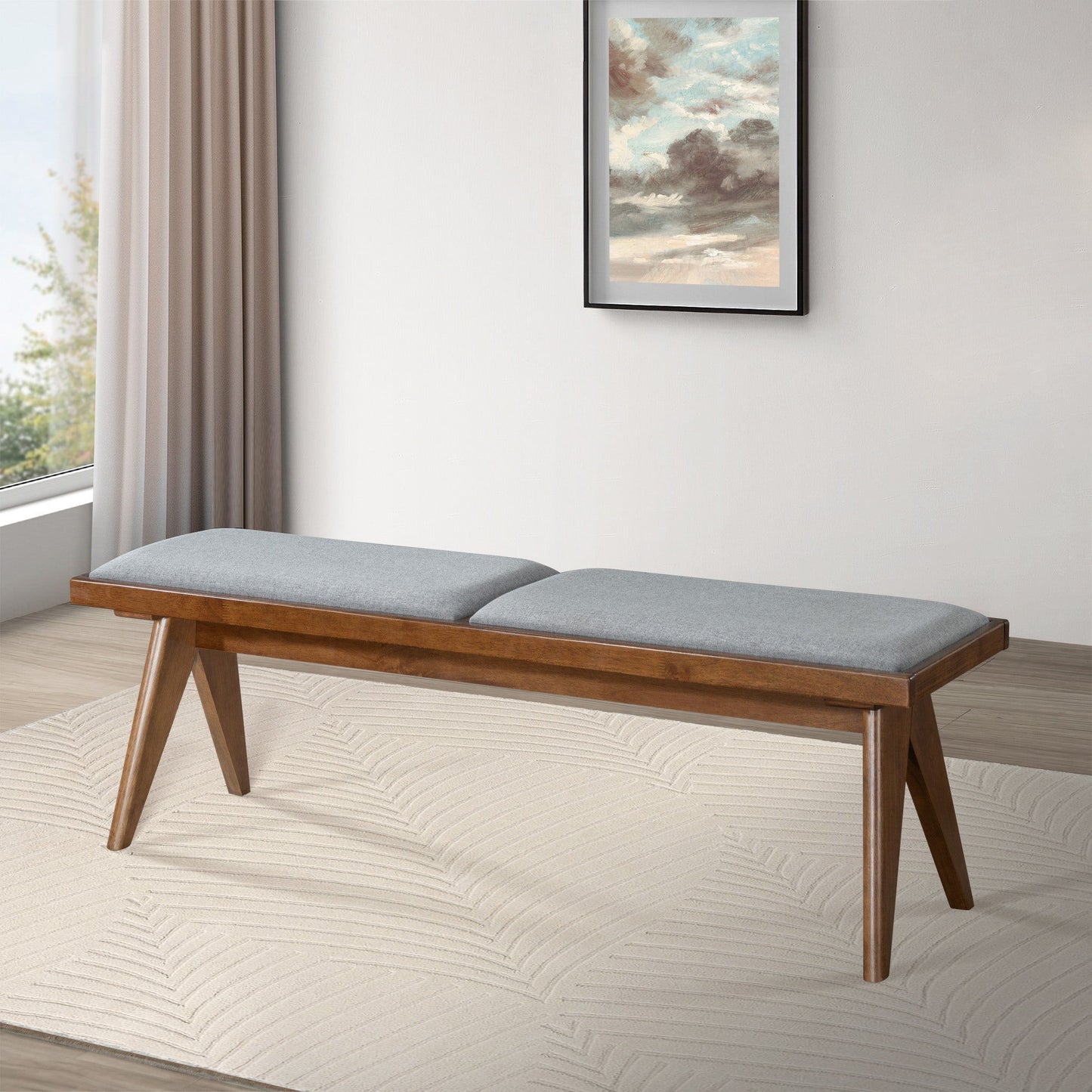 The Keira - Modern Aesthetics Bench, featuring angled wooden legs and two gray cushions, rests on a textured white rug in a modern room with light walls, sheer curtained window, and a framed painting.