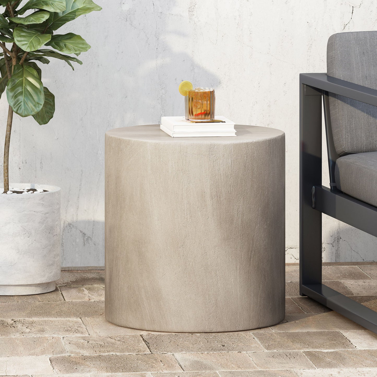 A Cylindrical Design, Outdoor Lightweight Concrete Side Table in beige is on a brick patio beside a gray outdoor sofa. On the table sit a drink with lemon and ice, and a white book. Nearby is a potted plant against a white wall.