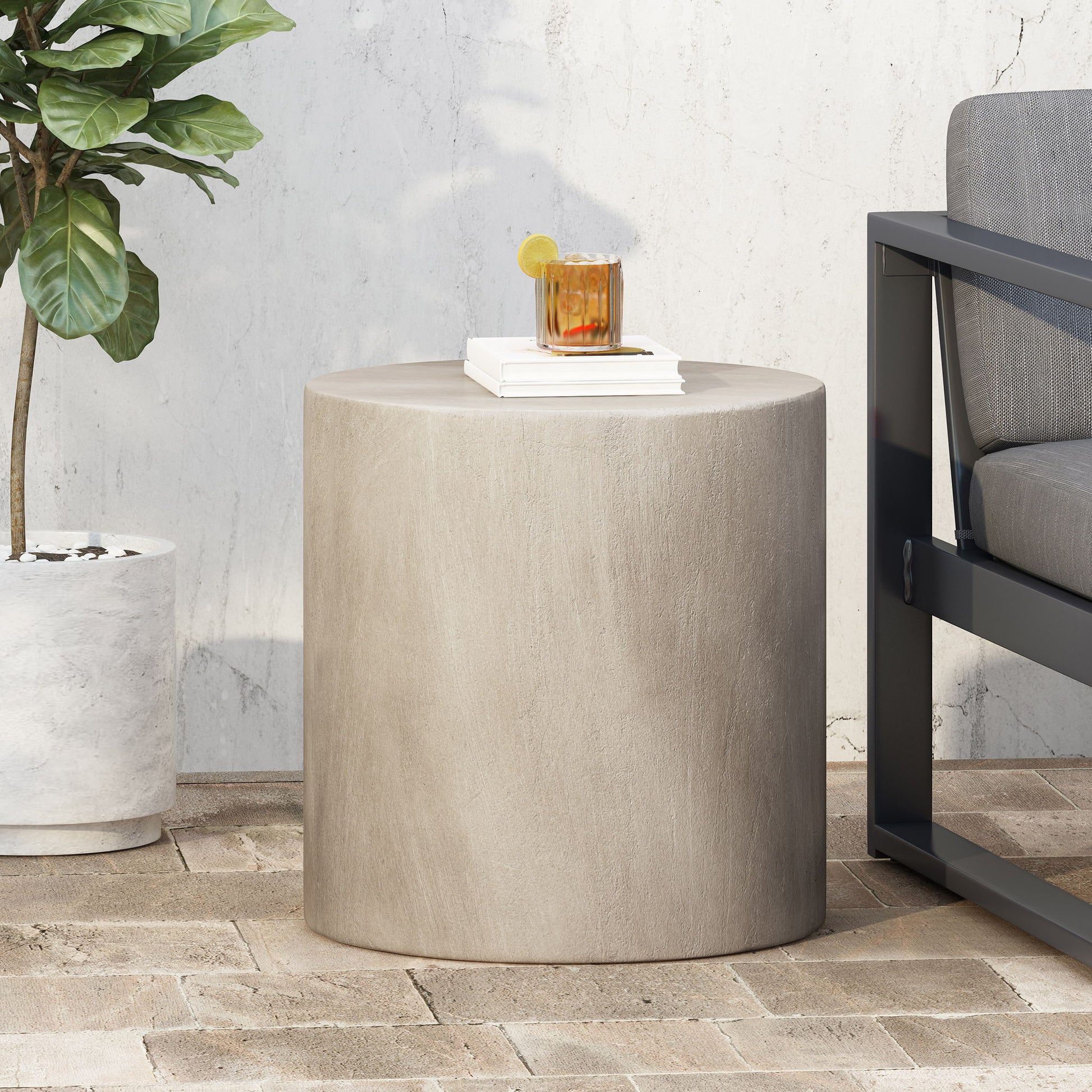 A Cylindrical Design, Outdoor Lightweight Concrete Side Table in beige is on a brick patio beside a gray outdoor sofa. On the table sit a drink with lemon and ice, and a white book. Nearby is a potted plant against a white wall.
