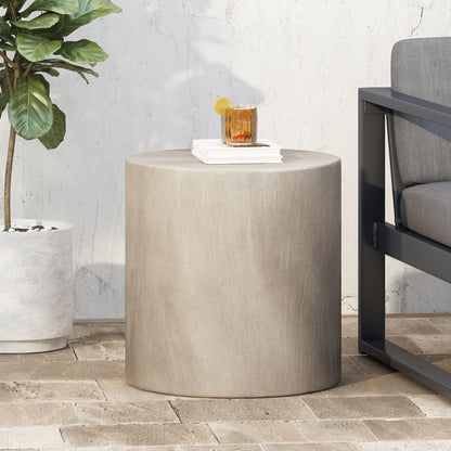 A Cylindrical Design, Outdoor Lightweight Concrete Side Table in beige is on a brick patio beside a gray outdoor sofa. On the table sit a drink with lemon and ice, and a white book. Nearby is a potted plant against a white wall.