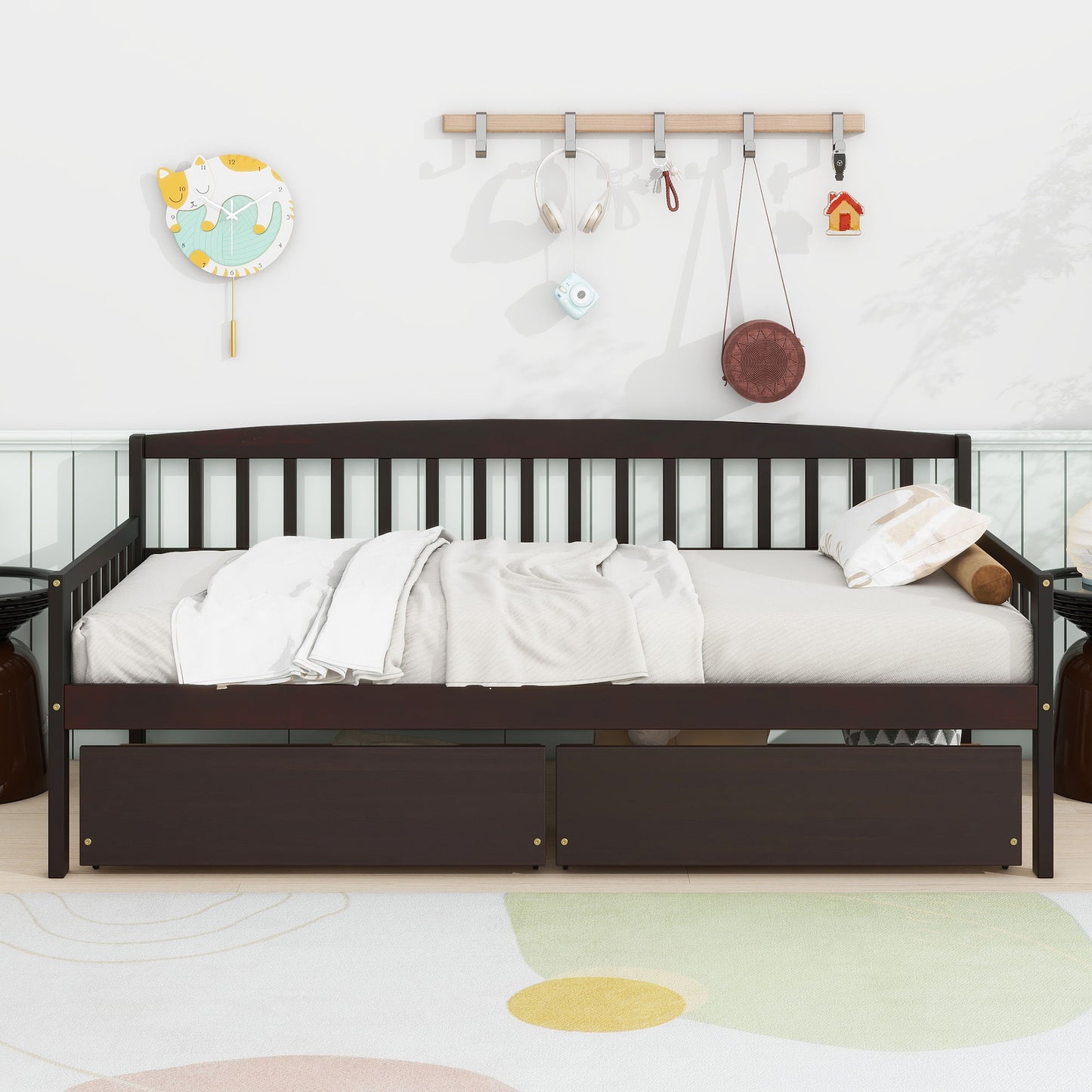 A Pine Wood Daybed With Two Storage Drawers, featuring a 10-slat bed platform, is neatly made with light bedding and pillows. Wall hooks above hold headphones and decorations, alongside a playful clock and wall art.