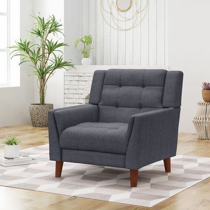A modern living room featuring the Luminaesque Fabric Upholstered Armchair with a tufted back in dark gray, placed on a light patterned rug with wooden floors, tall potted plants, wicker décor, and sunlight through large windows.