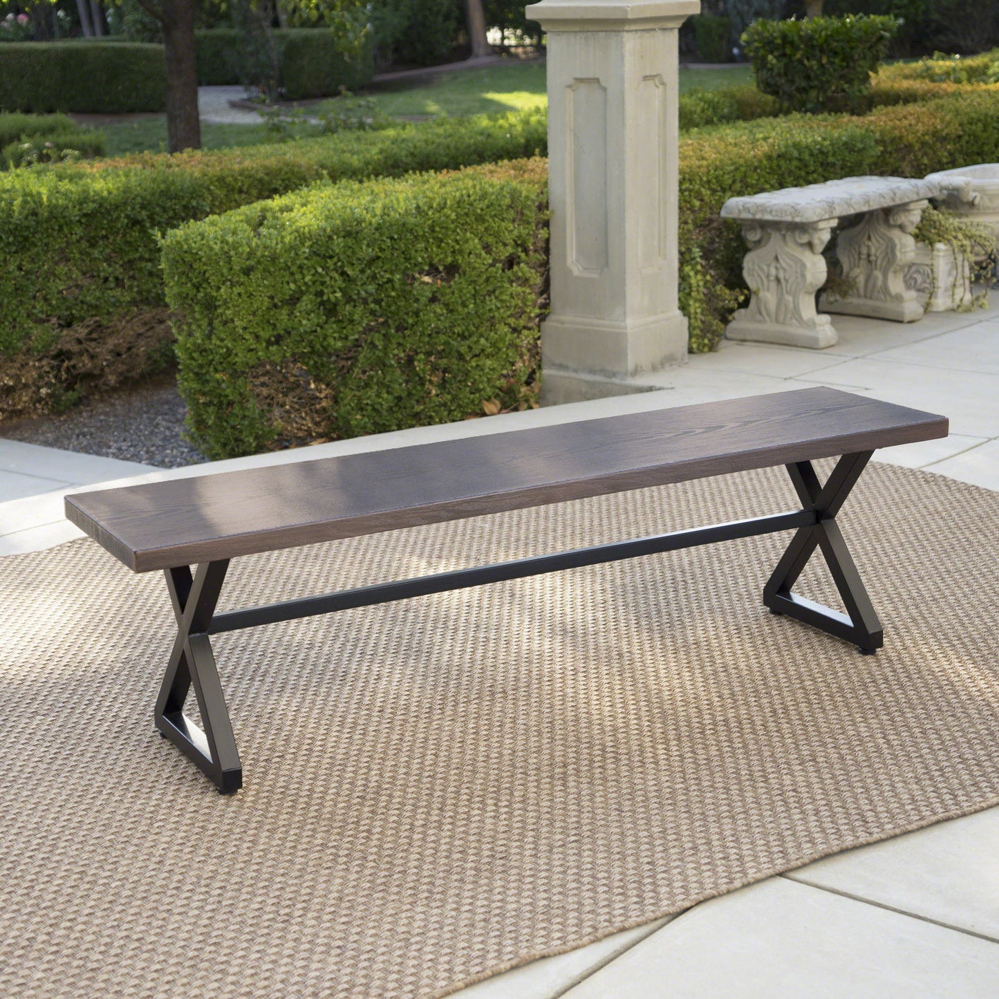 A Modern Aluminum Outdoor Dining Bench With X Base sits on a beige outdoor rug on a patio, surrounded by green hedges, stone benches, and trees in a garden setting.