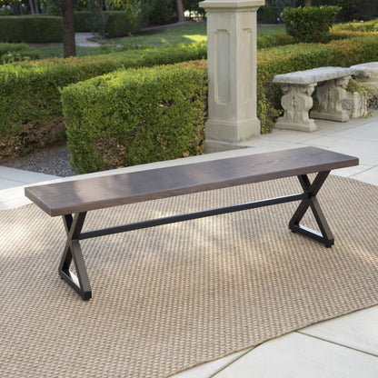 A Modern Aluminum Outdoor Dining Bench With X Base sits on a beige outdoor rug on a patio, surrounded by green hedges, stone benches, and trees in a garden setting.