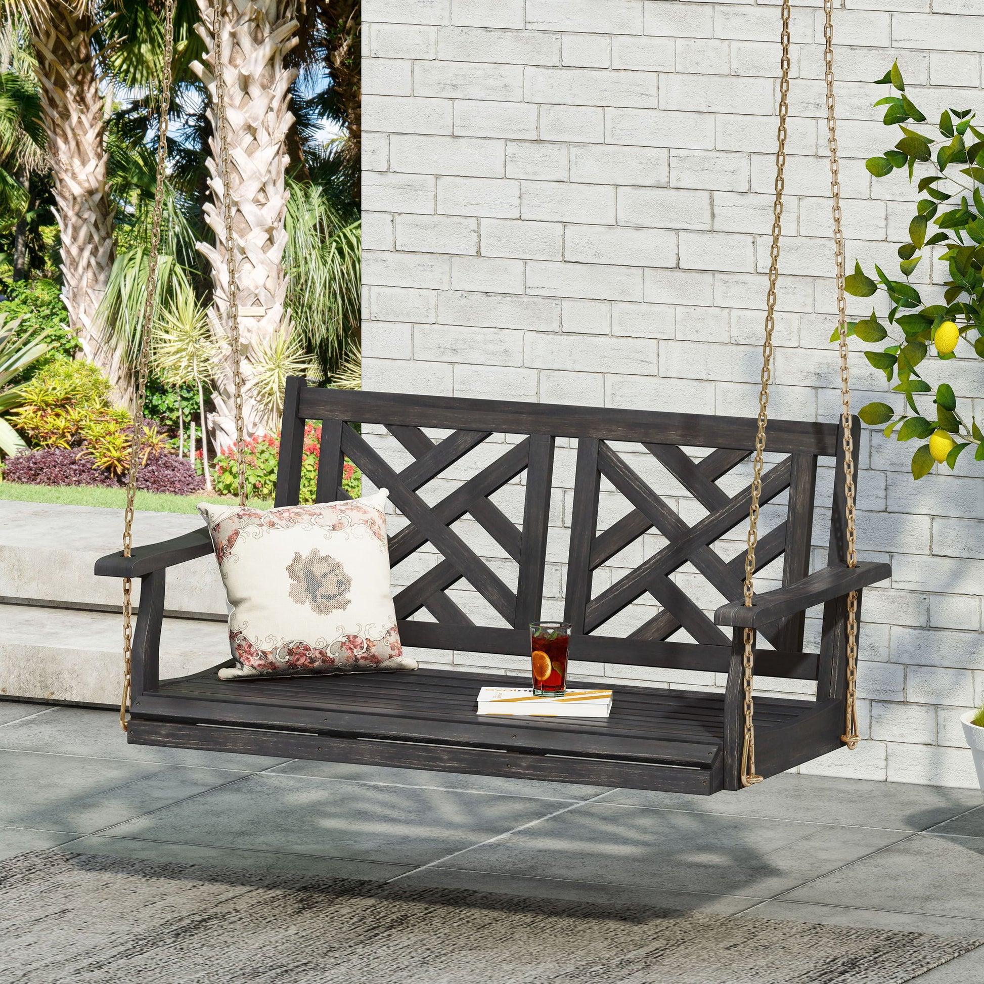 The Alejandro - Acacia Wood Porch Swing With Slat Panel Seating hangs from gold chains near a white brick wall, featuring a floral cushion and a glass of iced tea. Palm trees and lush green plants can be seen in the background.