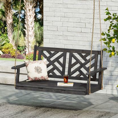 The Alejandro - Acacia Wood Porch Swing With Slat Panel Seating hangs from gold chains near a white brick wall, featuring a floral cushion and a glass of iced tea. Palm trees and lush green plants can be seen in the background.