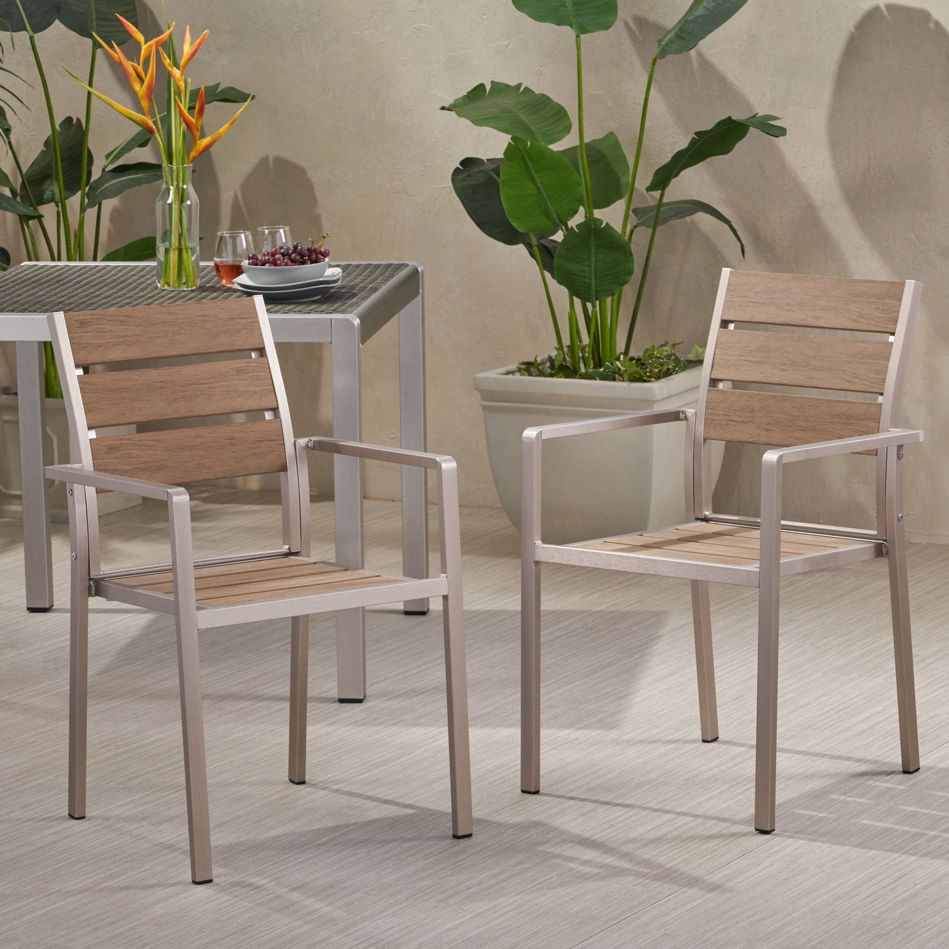 Two Dreamlan Outdoor Aluminum Dining Chairs (Set of 2) with wood slats are on a patio. A table behind holds drinks and fruit, while large green potted plants sit against a textured wall.