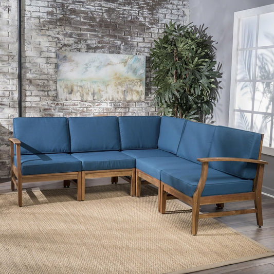 The Perla - 5 Pieces Sofa Set, a modern L-shaped wooden sofa with blue cushions, sits on a light woven rug in a bright room featuring a brick wall, large potted plant, landscape artwork, and sunlight streaming through the window.