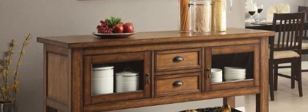 A Kadri Kitchen Cart in Distressed Chestnut stands with drawers and glass-front cabinets displaying jars, plates, cookware, and a fruit bowl. A dining table and chairs appear in the background.