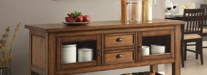 A Kadri Kitchen Cart in Distressed Chestnut stands with drawers and glass-front cabinets displaying jars, plates, cookware, and a fruit bowl. A dining table and chairs appear in the background.