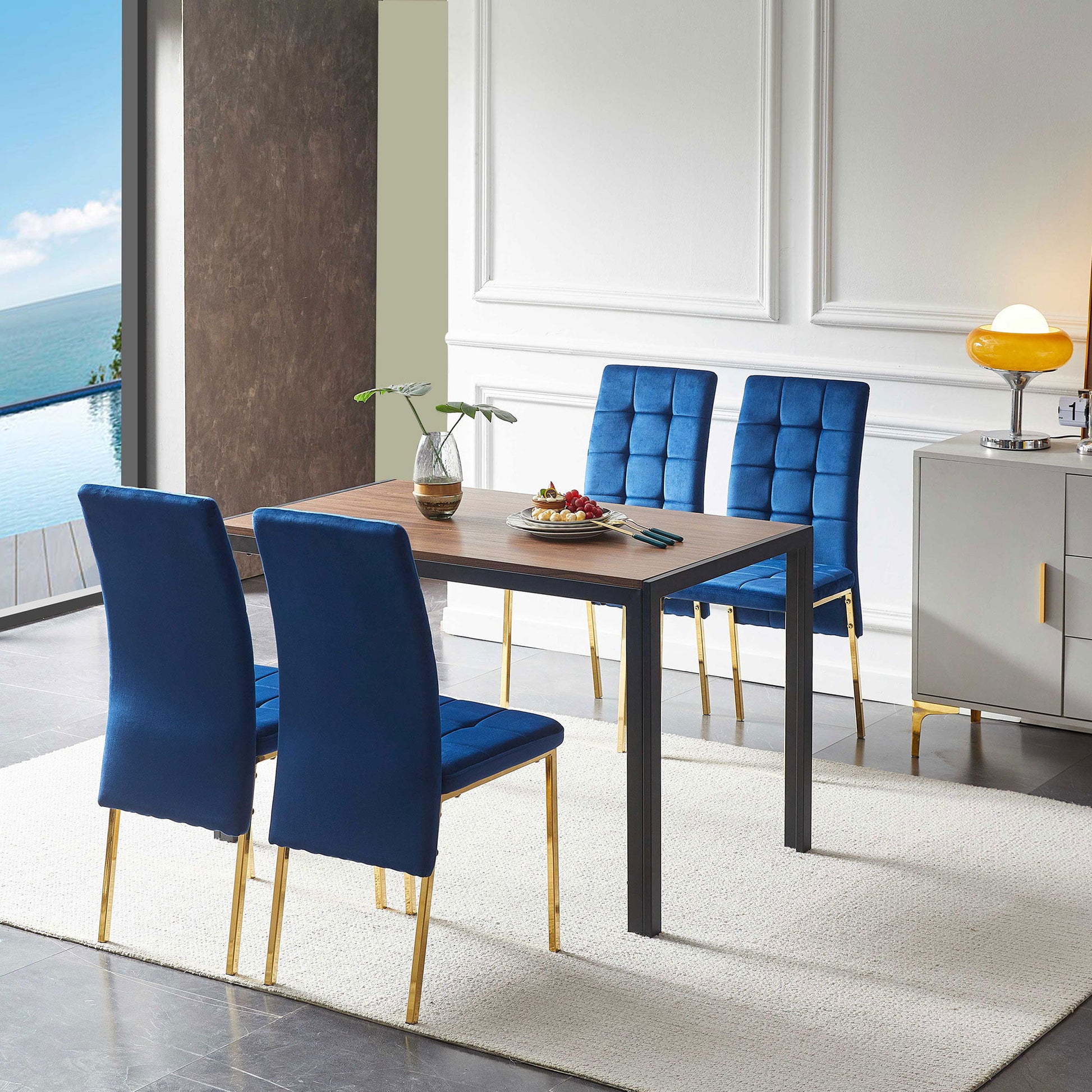 A modern dining room features a 5-piece set with velvet high-back Nordic chairs with golden legs and a creative dining table, complemented by a white rug, potted plant, fruit plate, lamp on sideboard, and ocean view through an open sliding door.