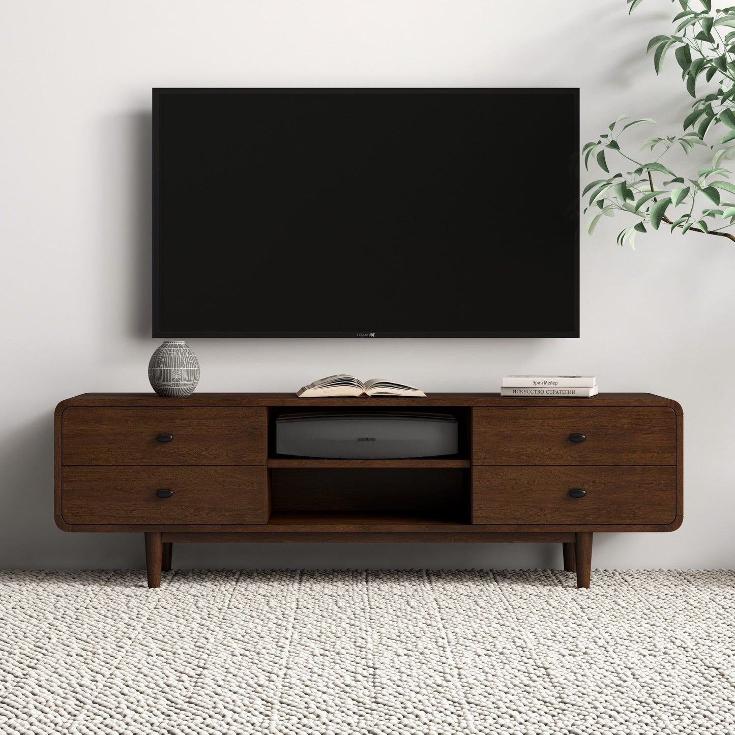 A modern living room features a flat-screen TV mounted above the Alexa Mid-Century Modern Style TV Stand in dark brown wood, which holds a vase, books, an electronic device, and is complemented by a plant in the corner.